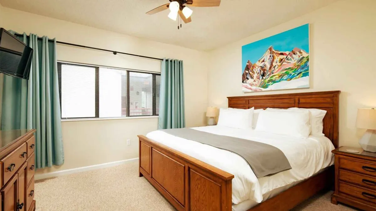 A serene bedroom featuring a plush king-sized bed with crisp linens, inviting pillows, and soft lighting. Artwork adorns the wall above the headboard, while blackout curtains frame a large window, creating an intimate, restful atmosphere. A sturdy dresser sits opposite the bed, and the space is imbued with a sense of calm and privacy.