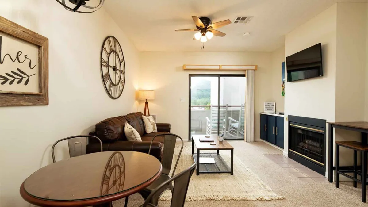 The image shows a cozy, stylishly decorated condo interior with a plush sofa adorned with pillows, a dining table set for gatherings, and a crackling fireplace adjacent to a modern television. The setup invites relaxation, socializing, and enjoying quiet moments, all within a warm, inviting atmosphere.