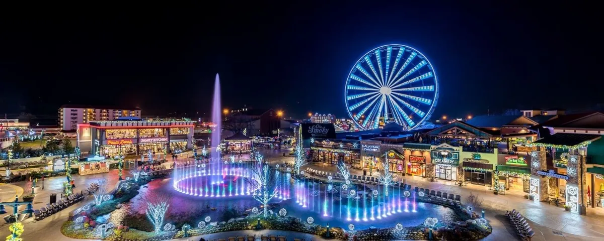 A lively nighttime scene featuring a fountain illuminated with colorful lights, a giant Ferris wheel, and a collection of inviting shops and restaurants bustling with activity. The area exudes vibrancy and excitement, ideal for evening strolls or romantic outings in downtown Gatlinburg.