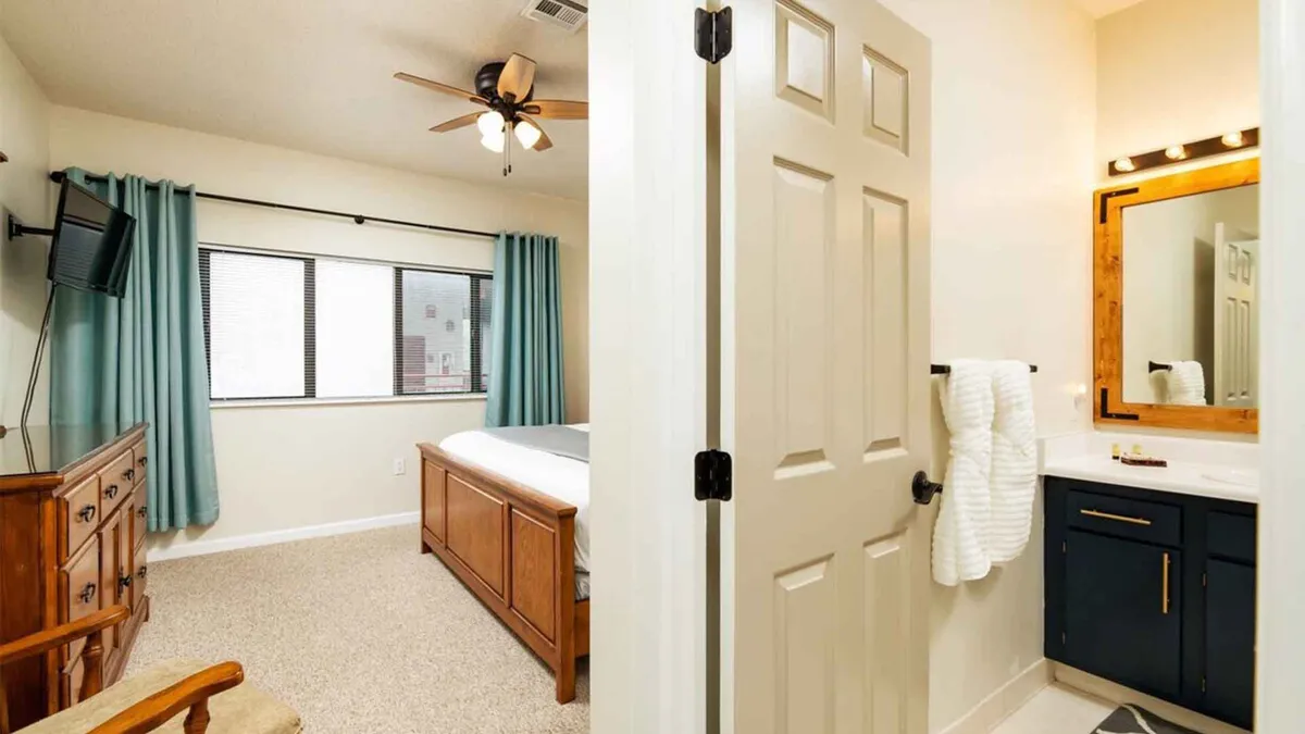 A cozy bedroom scene with the bed taking center stage, positioned to catch soft natural light from the window. A glimpse of the adjacent bathroom mirror hints at the suite-like arrangement, offering seamless transition between sleeping and bathing spaces. The room exudes warmth and an inviting, homey feel.