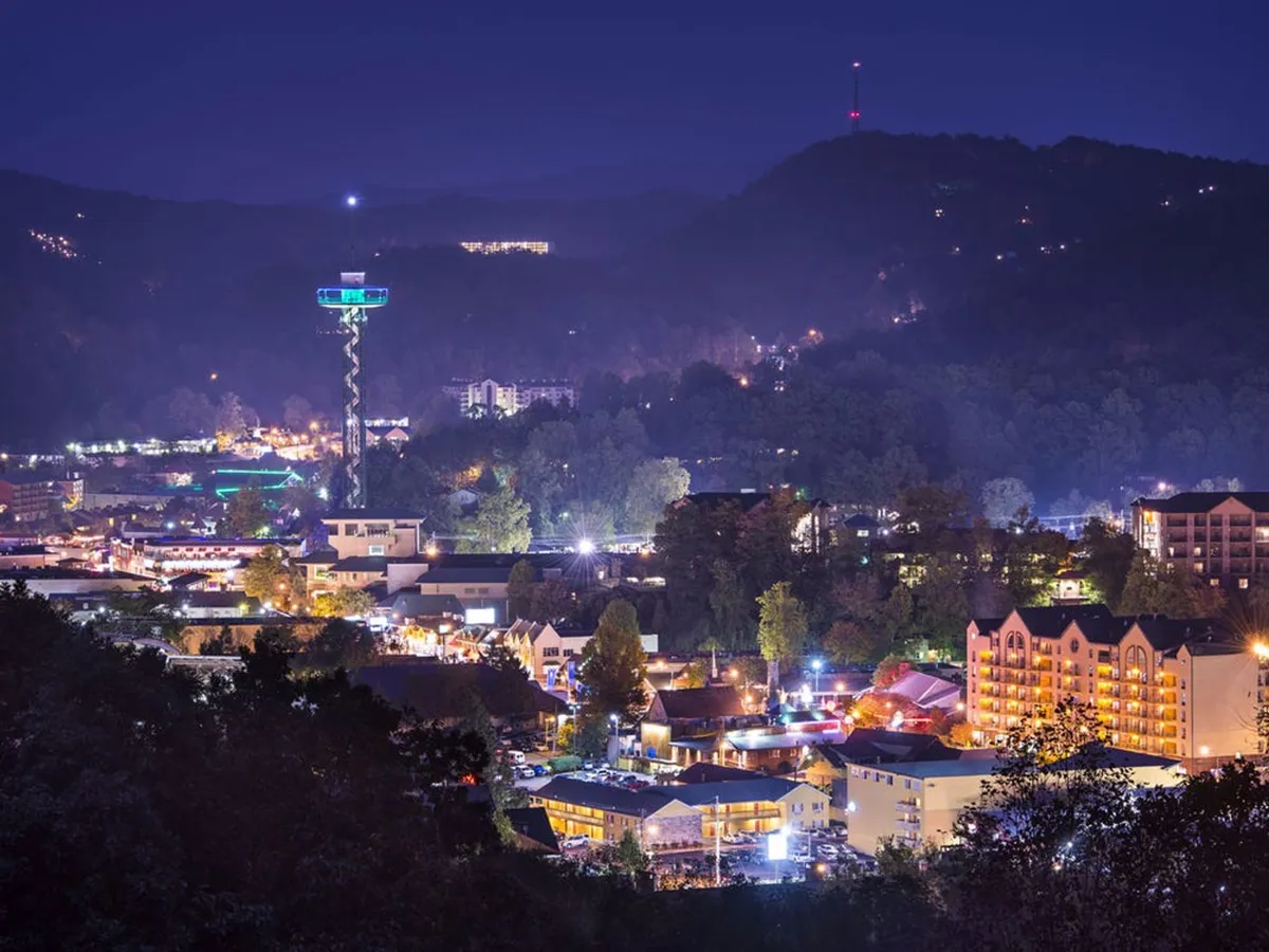 A vibrant nighttime cityscape with neon lights illuminating hotel buildings, a prominent tower structure (likely the Gatlinburg Space Needle), and the dramatic backdrop of nearby mountains. The scene conveys the excitement and accessibility of downtown attractions.