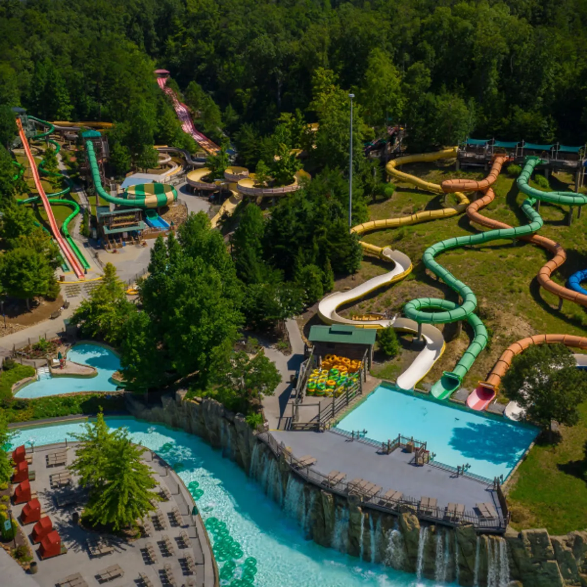 A lively outdoor scene featuring multiple water slides cascading into resort-style swimming pools, surrounded by lush greenery and deck chairs. Families and groups can be seen enjoying the water, sunbathing, and creating joyful memories together.