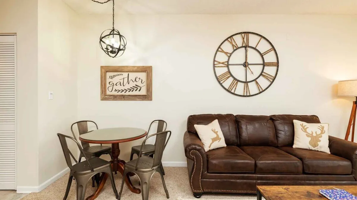 An inviting dining area featuring a brown leather sofa adjacent to a round dining table, accented by a stylish wall clock and a 'gather' sign. The room radiates warmth and is set up for dining, socializing, and sharing meals with loved ones, reinforcing the idea of connection and togetherness.
