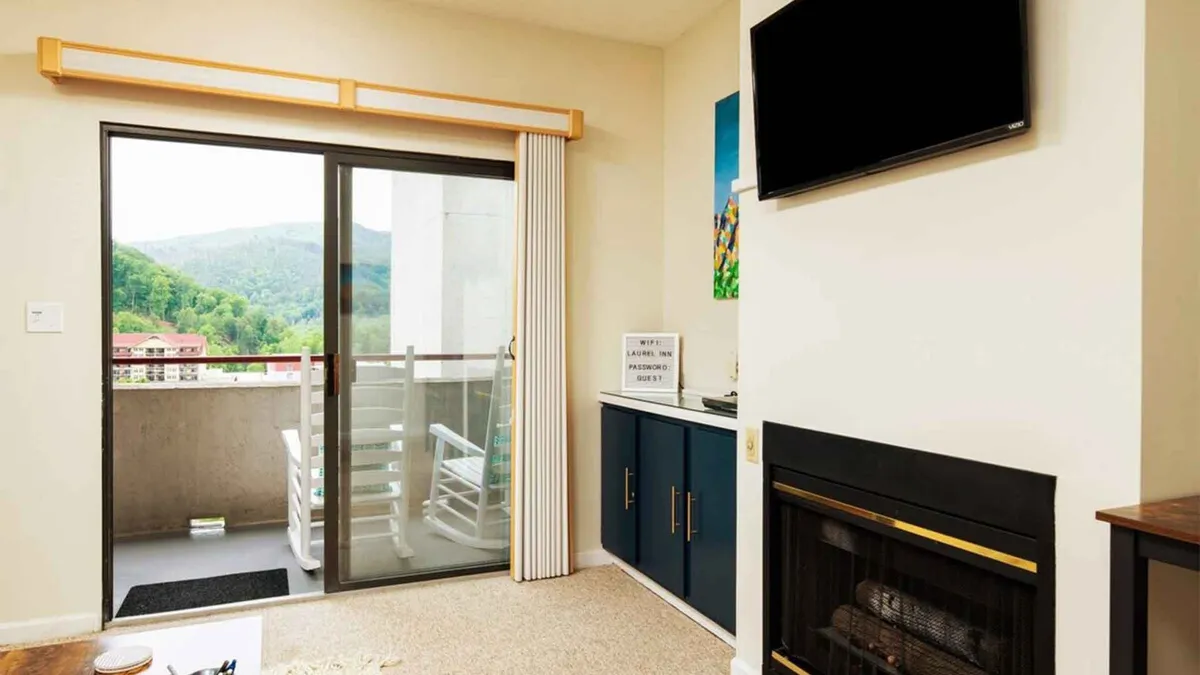 A bright, inviting living area featuring a cozy fireplace nestled into the wall, a large television, and a glass door opening directly onto a private balcony. Through the door, you can glimpse mountain scenery, while the contemporary furnishings and tranquil setting evoke comfort and relaxation.
