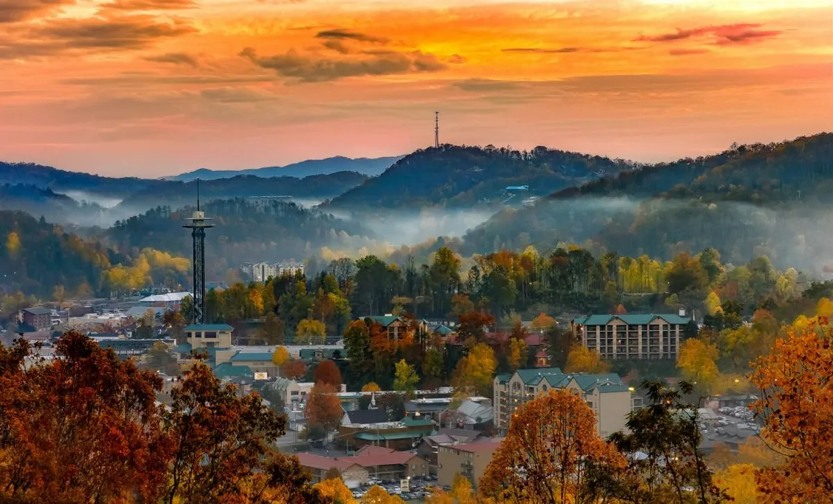 A panoramic sunset view over Gatlinburg, with mountains framing the charming town buildings and autumn foliage adding warmth and color. The sky is painted in soft golden hues, creating a serene and breathtaking backdrop.