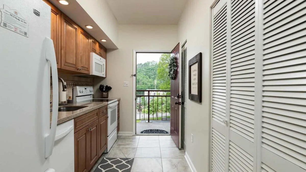 A welcoming interior scene showing the kitchen area that opens toward a door leading to a balcony. The decorative wall art and inviting cabinetry suggest a space for gathering and entertaining, while the view through the doorway hints at the outdoors beyond.