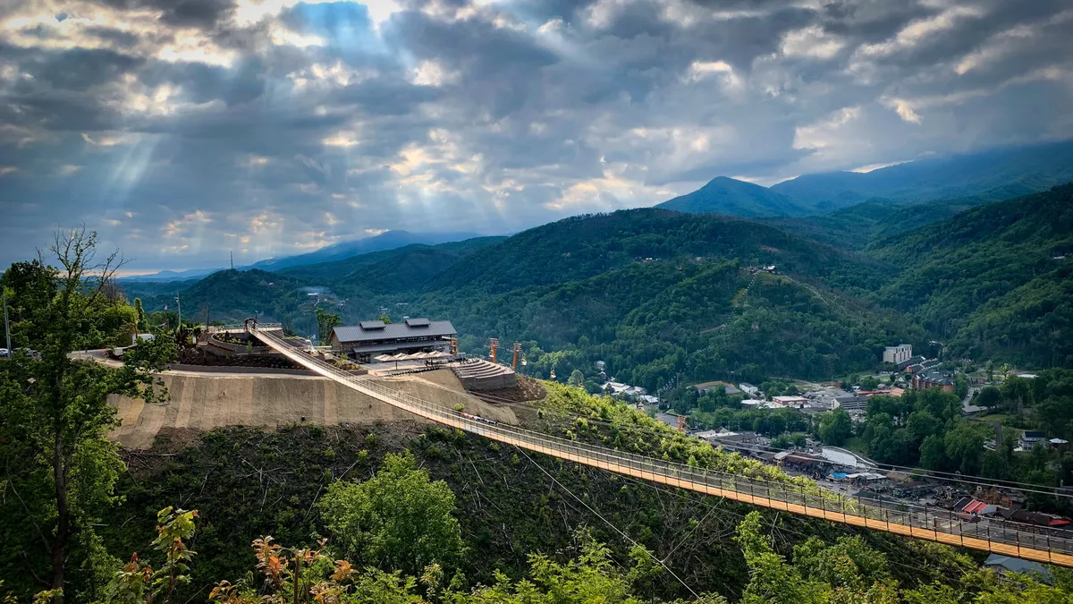 A sweeping view of a mountainous landscape with a striking suspension bridge crossing a lush valley. A modern building structure is visible, and the setting invites outdoor activities such as hiking, zip-lining, and photography. The scene is invigorating, capturing both the grandeur of the Smokies and the thrill of exploration.