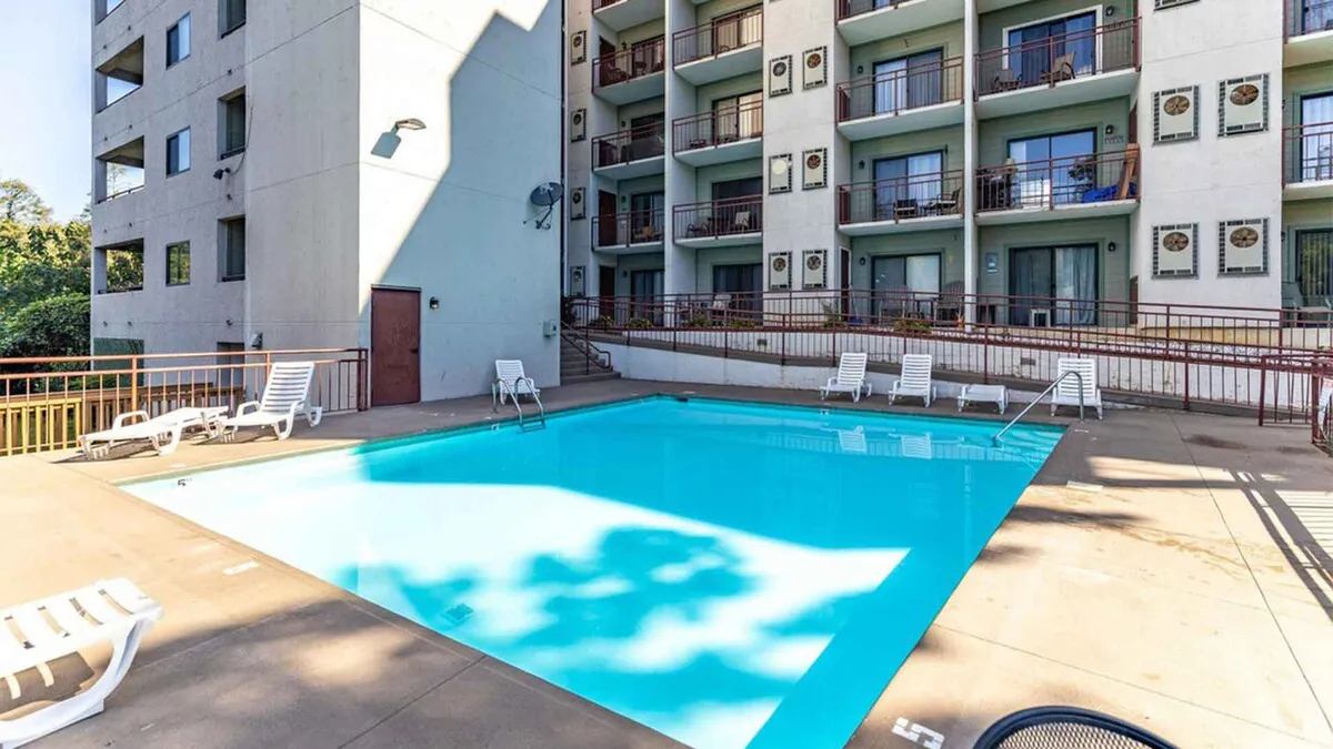 A vibrant exterior scene of a swimming pool with inviting lounge chairs, the building's facade in the background, and a relaxing mood. People are shown swimming, sunbathing, and enjoying the pool area, suggesting both activity and relaxation options.